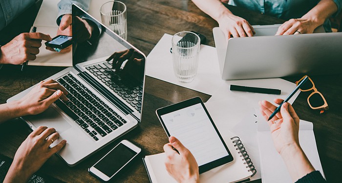 Laptops, mobile phones, tablets and documents, with users hands showing, on a working table.
